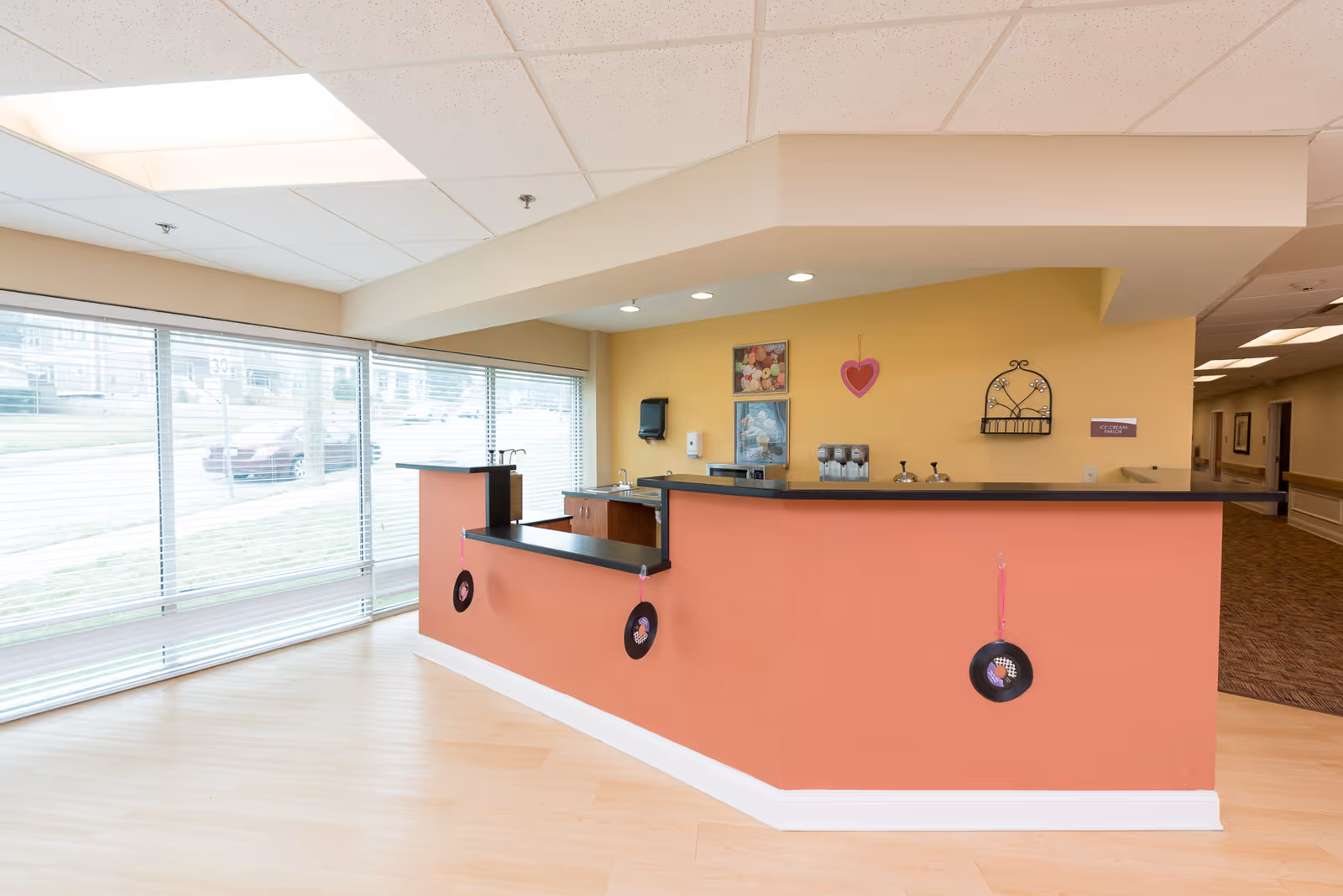 Reception desk area inside a senior living facility with large windows covered by horizontal blinds, a peach-colored front desk decorated with hanging vinyl records, and a yellow wall behind the desk featuring a heart decoration, framed pictures, and a small shelf. The floor is light wood, and there is a hallway visible to the right.
