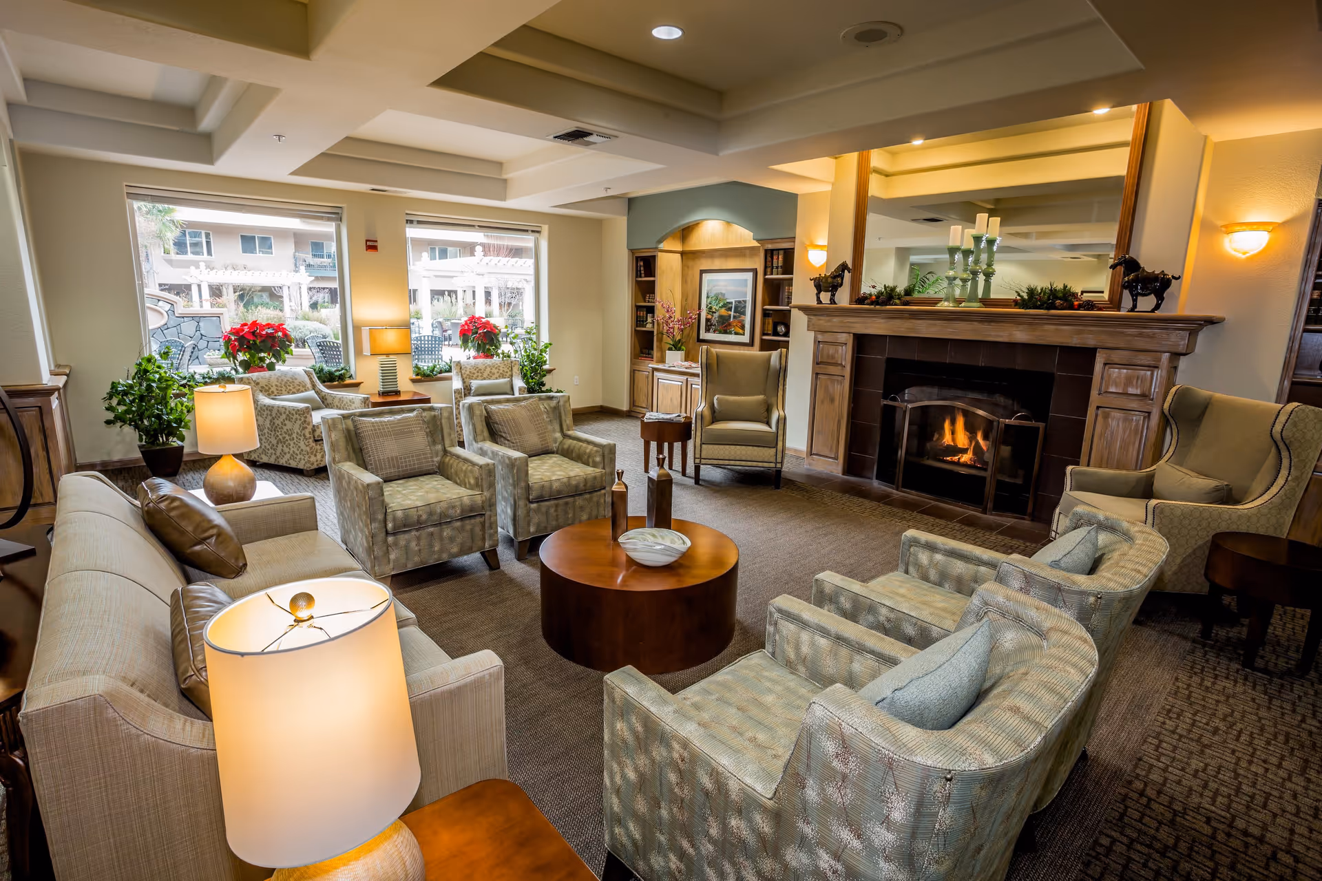 A cozy living room area with multiple upholstered armchairs and sofas arranged around a round wooden coffee table. The room features a lit fireplace with a large mirror above it, decorative items on the mantel, and built-in wooden shelves with books and artwork. Large windows let in natural light and show an outdoor patio area with chairs and plants.