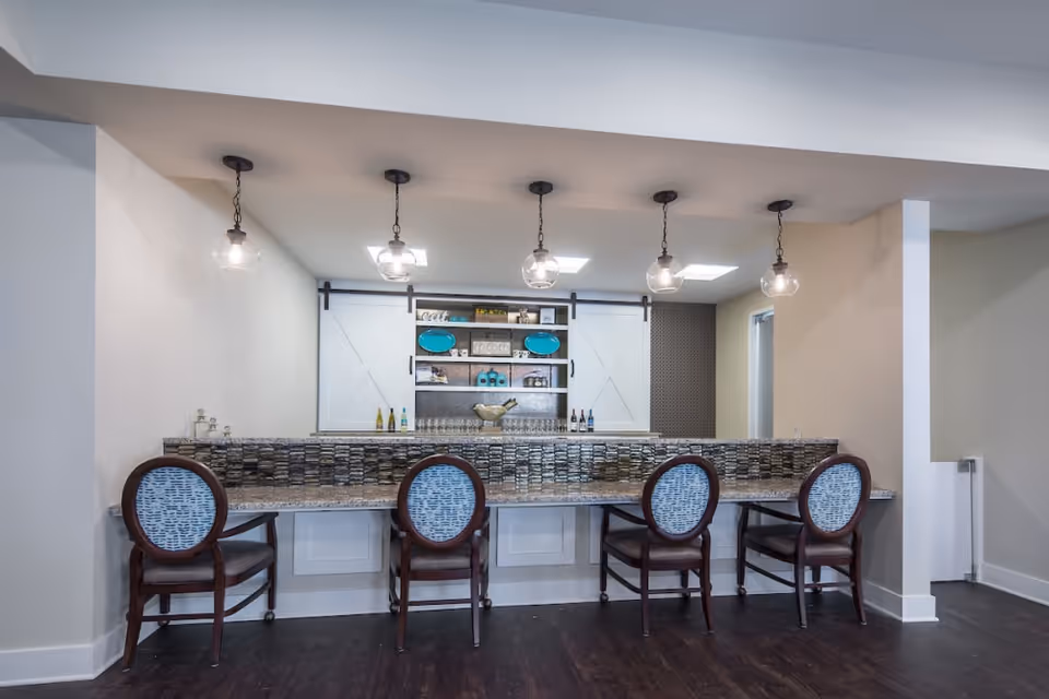 Communal bar-style counter with four upholstered chairs, pendant lights, and decorative shelving with bottles behind the counter.