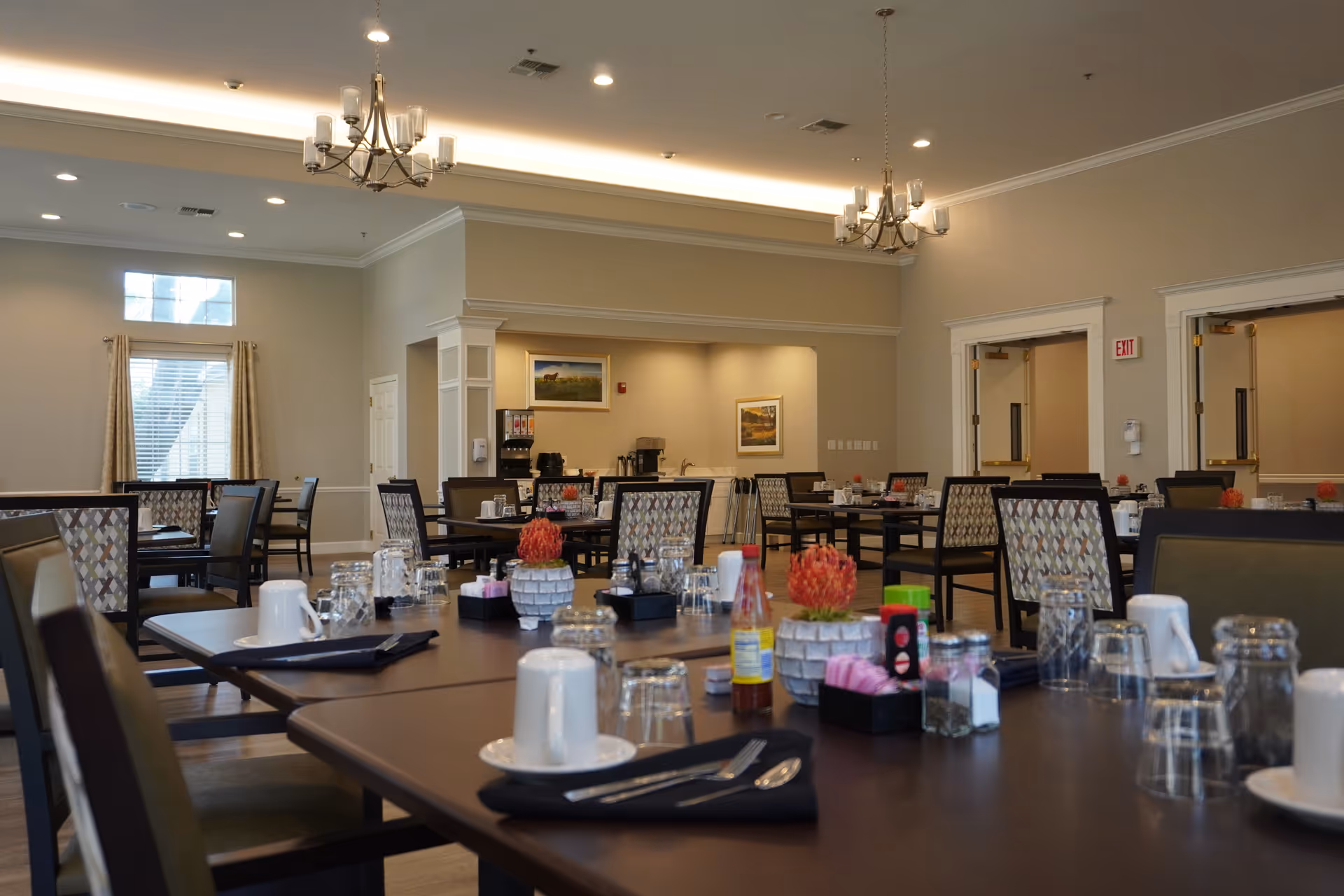 A spacious dining room with multiple tables and chairs arranged neatly. Each table is set with white coffee cups, glasses, silverware on black napkins, and small decorative flower arrangements. The room has beige walls, two chandeliers hanging from the ceiling, and large windows with curtains allowing natural light. In the background, there is a beverage station with coffee dispensers and framed artwork on the walls.