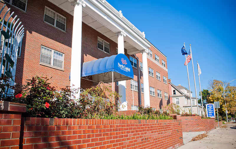 Front exterior of a red-brick nursing and rehabilitation building with white columns, a blue awning labeled 'North Capital', flags, and a low brick wall.