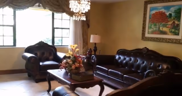 A cozy living room with dark leather tufted sofas and armchairs arranged around a wooden coffee table adorned with a floral centerpiece. A chandelier hangs from the ceiling, and a framed painting of a house with trees is on the wall. A table lamp is placed on a side table near the sofa, and large windows with curtains allow natural light into the room.