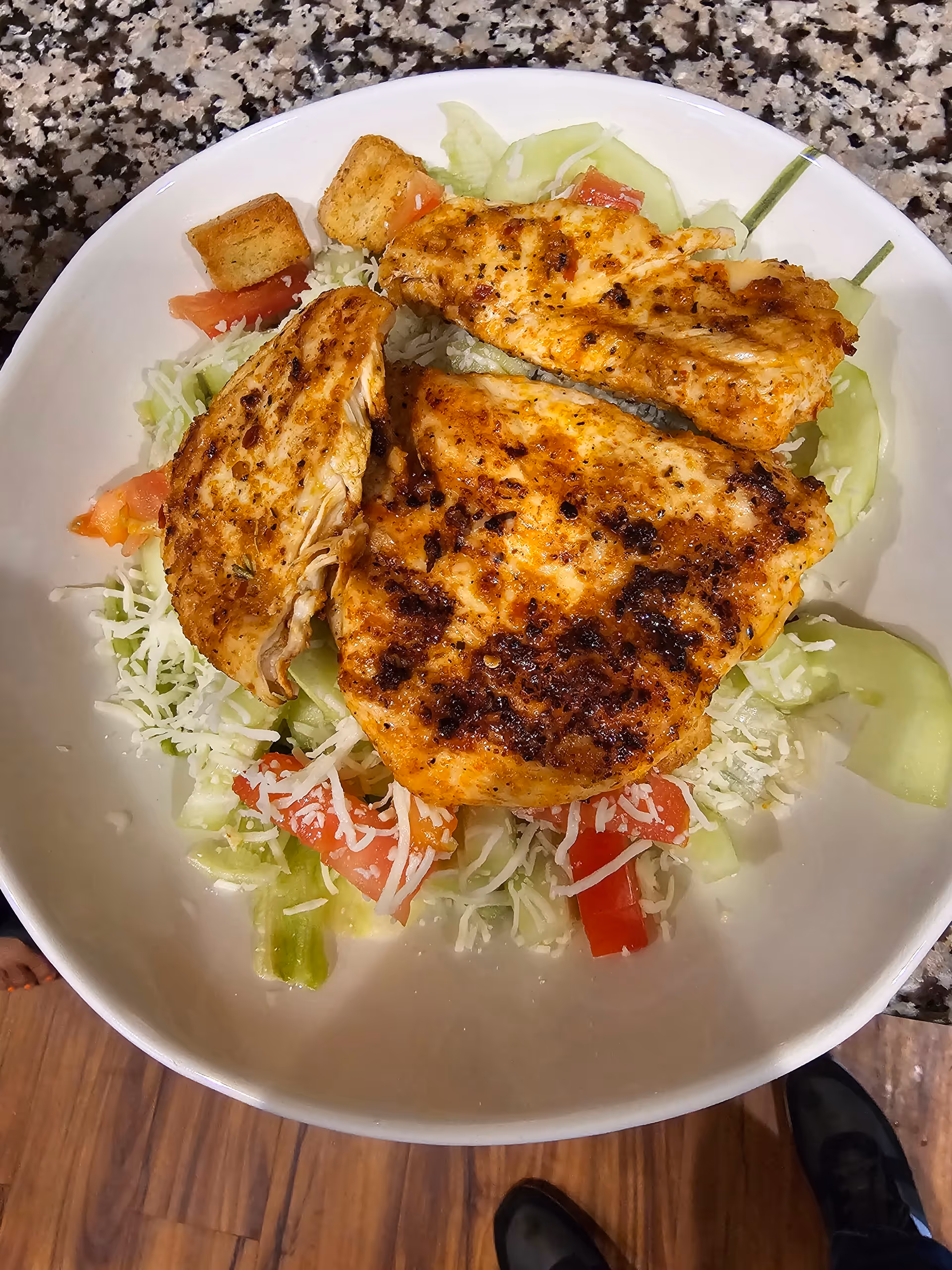 A white bowl containing a salad with shredded lettuce, diced tomatoes, cucumber slices, shredded cheese, croutons, and three pieces of grilled chicken on top. The bowl is placed on a speckled countertop with a wooden floor visible below.