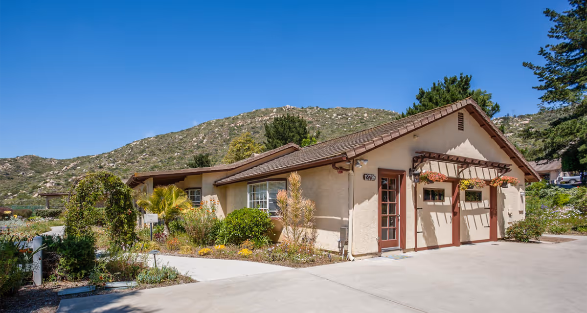 Exterior view of a single-story building with beige walls and a brown tiled roof at Poway Gardens Senior Living - Mountain Vistas, surrounded by landscaped gardens and a mountainous backdrop under a clear blue sky.