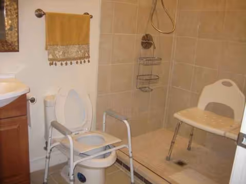 Accessible bathroom with a raised toilet, shower chair, grab bars, and tiled walk-in shower.
