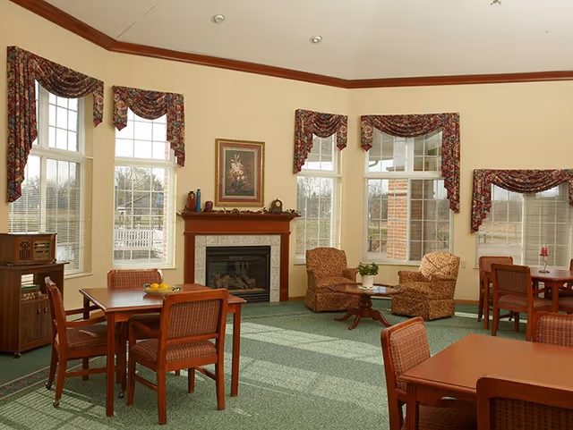 A bright, spacious common area with large windows adorned with floral valances. The room features a fireplace with a framed floral painting above it, two upholstered armchairs around a small wooden coffee table, and several wooden tables with matching chairs. The carpet is green, and the walls are painted a light cream color with a wooden trim near the ceiling.