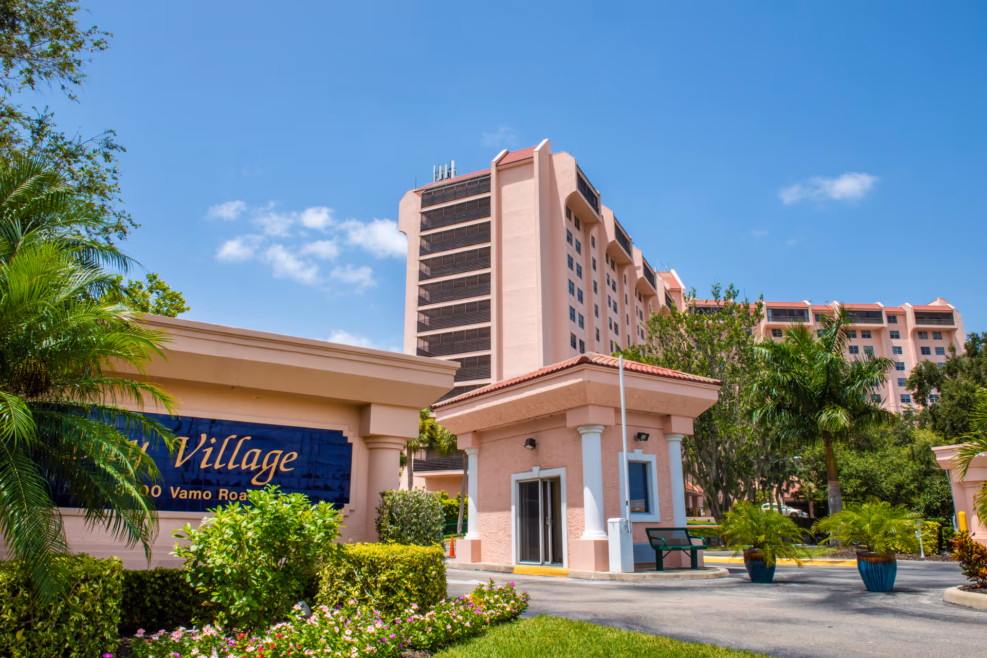 Exterior view of Bay Village of Sarasota Inc, showing a multi-story pink building with balconies, a guardhouse at the entrance, lush green landscaping, palm trees, and a clear blue sky.