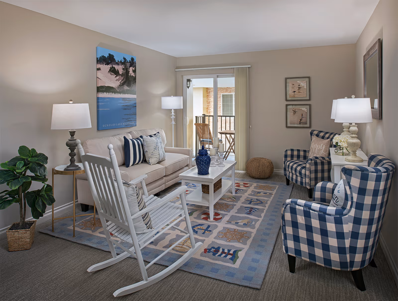 A cozy living room with a beige sofa adorned with striped and patterned pillows, a white wooden rocking chair with a cushion, and two blue and white checkered armchairs. A white coffee table with decorative items sits on a patterned rug with nautical designs. The room has beige walls, two table lamps, a potted plant, framed artwork, and a sliding glass door leading to a small balcony with outdoor furniture.