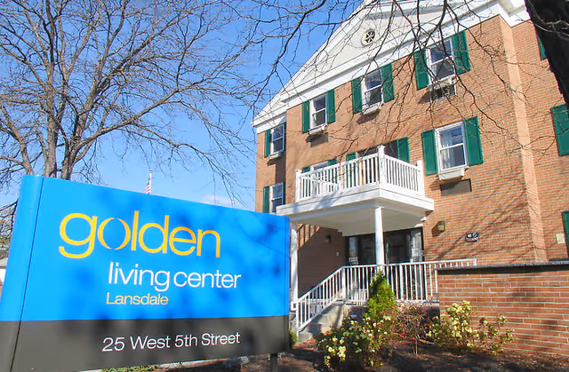 Exterior view of a brick building with green window shutters and a white balcony. In front of the building is a blue and black sign that reads 'golden living center Lansdale 25 West 5th Street'. Leafless trees are visible against a clear blue sky.