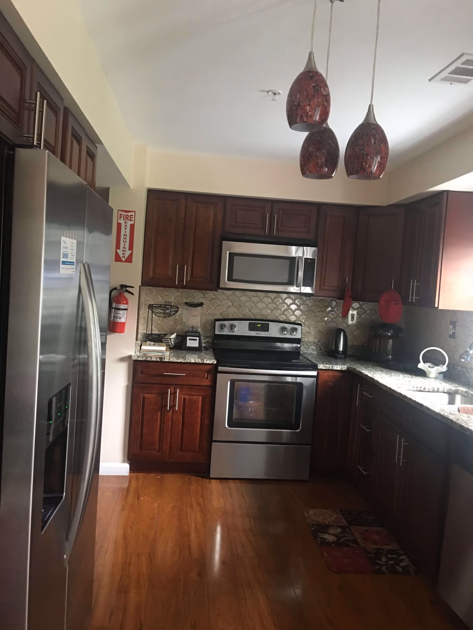A kitchen with wooden cabinets, stainless steel refrigerator, oven, and microwave. The countertops are granite with various kitchen appliances including a blender, kettle, and coffee maker. Three pendant lights with red shades hang from the ceiling. A fire extinguisher is mounted on the wall next to the refrigerator.
