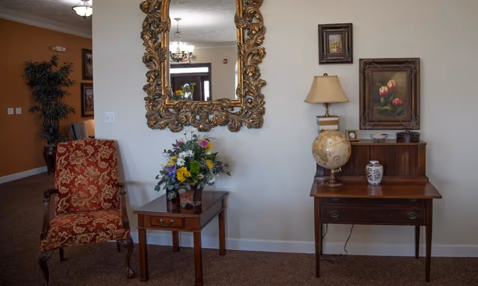 A cozy interior corner featuring a red patterned armchair next to a wooden side table with a colorful flower arrangement. Above the table hangs an ornate gold-framed mirror. To the right, there is a wooden desk with a globe, a lamp, a small framed picture, and decorative items. Two framed floral paintings are mounted on the wall above the desk.