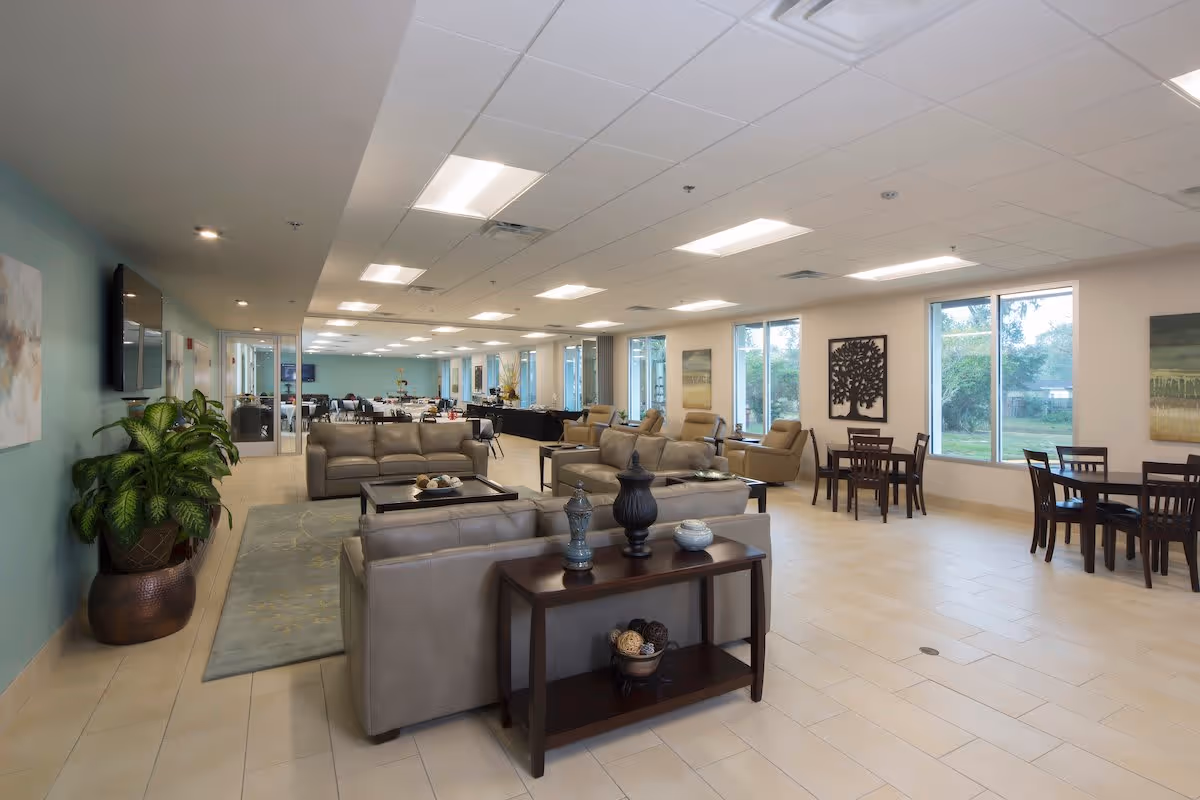 A spacious and well-lit senior living common area featuring multiple beige leather sofas and armchairs arranged around coffee tables. There are several wooden dining tables with chairs near large windows that let in natural light. The walls are decorated with artwork, and there is a large potted plant near a mounted flat-screen TV. The floor is tiled, and the ceiling has recessed lighting.