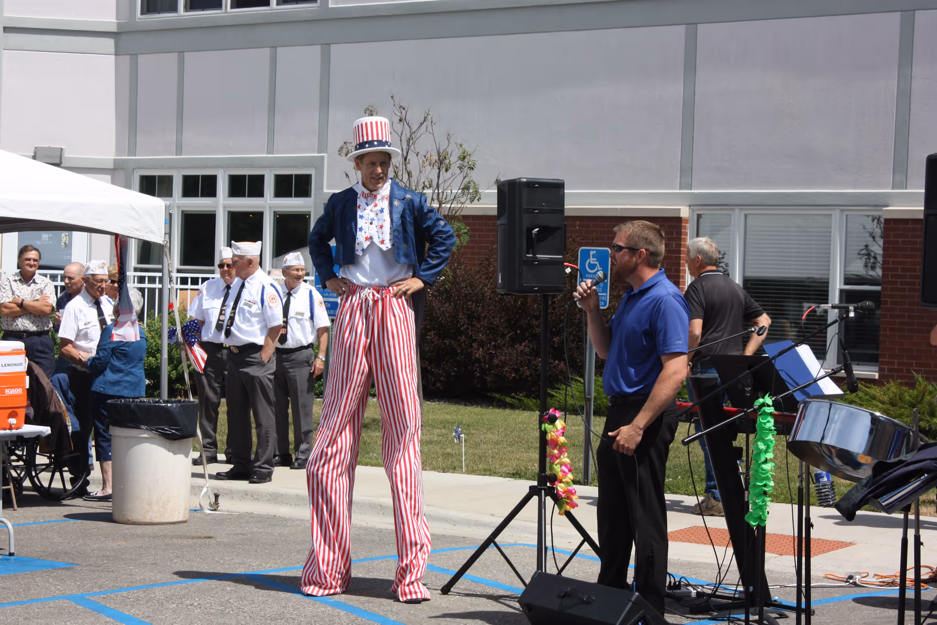 An outdoor event at PioneerCare Center with a man dressed in a patriotic Uncle Sam costume on stilts, standing next to a man speaking into a microphone. Several people, including veterans in uniform, are gathered in the background near a building. Musical instruments and speakers are set up nearby.