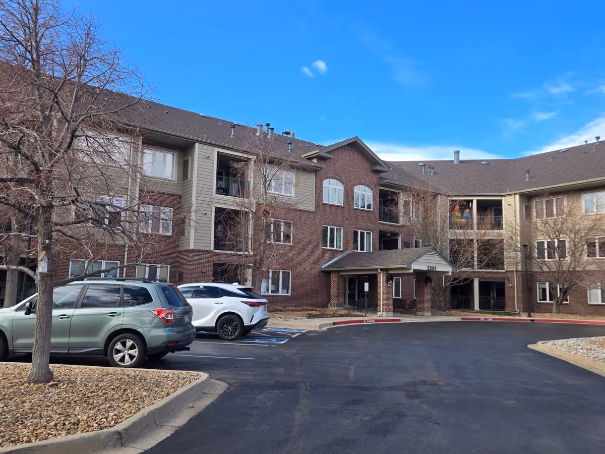 Front exterior of a three-story brick senior living building with parked cars, leafless trees, and a clear blue sky.
