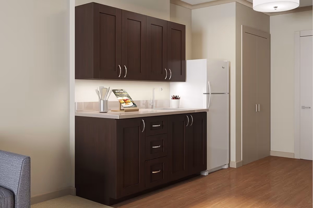 A small kitchen area with dark wood cabinets, a white countertop, a white refrigerator, and a sink. There is a utensil holder and a cookbook on the counter. The floor is wooden, and there is a gray couch partially visible on the left side.
