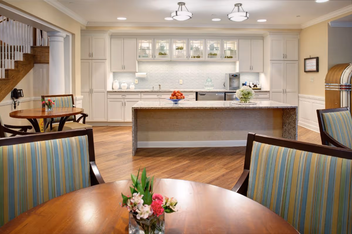 A bright and clean kitchen area with white cabinets, a large granite island in the center, and wooden floors. There are tables with striped upholstered chairs and small flower arrangements on the tables. A staircase with white railings and wooden banister is visible on the left side, and a vintage jukebox is on the right side of the image.