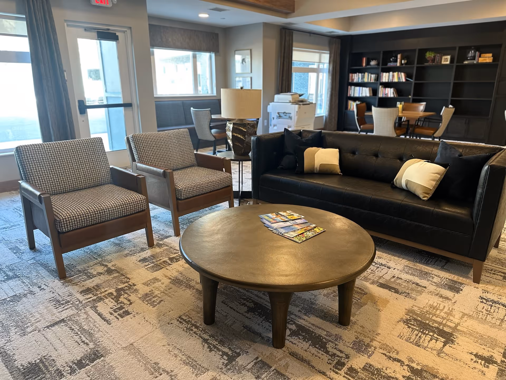 A cozy lounge area in a senior living facility featuring a black leather sofa with cushions, two patterned armchairs, a round wooden coffee table with magazines on it, a lamp on a side table, and a bookshelf with books and decorative items in the background. Large windows and a door let in natural light.