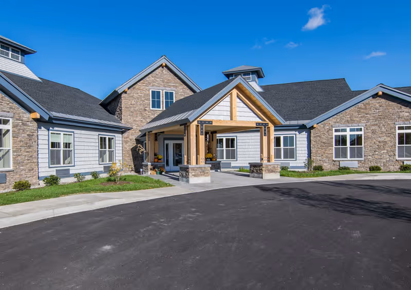 Exterior view of a senior living facility building with a covered entrance supported by wooden beams and stone pillars. The building has a combination of stone and light gray siding with multiple windows under a clear blue sky.