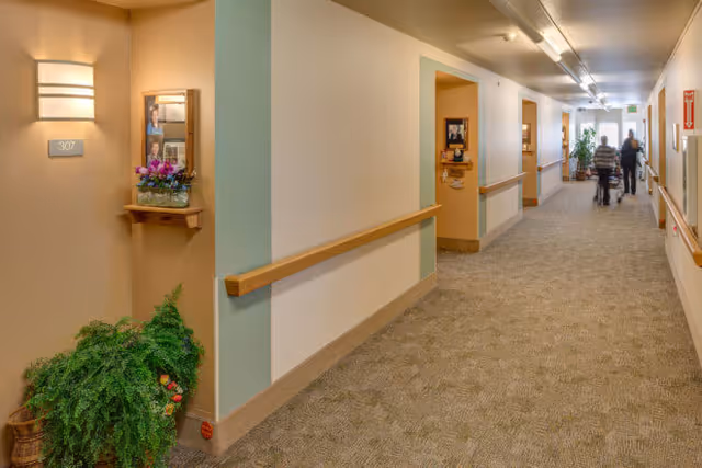 A well-lit hallway in an assisted living facility with beige walls and carpeted floor. The hallway has wooden handrails on both sides and several doorways leading to rooms. A small shelf with a flower arrangement and framed photos is mounted on the left wall near room 307. Two people are walking down the hallway in the distance.