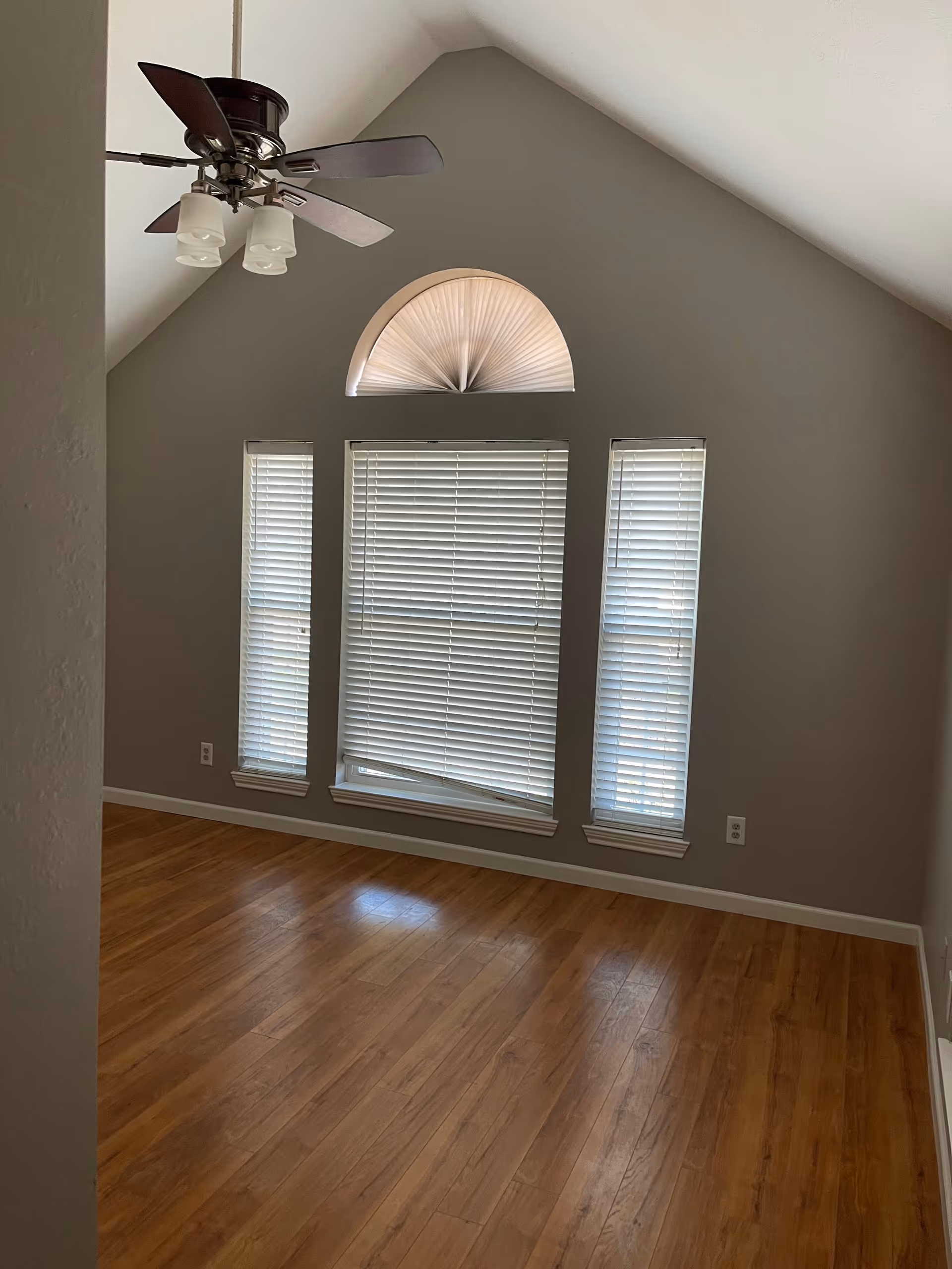 Empty room with wooden flooring, three rectangular windows with closed blinds, and a semi-circular window with a pleated shade above them. A ceiling fan with four wooden blades and three light fixtures hangs from the vaulted ceiling. The walls are painted gray.