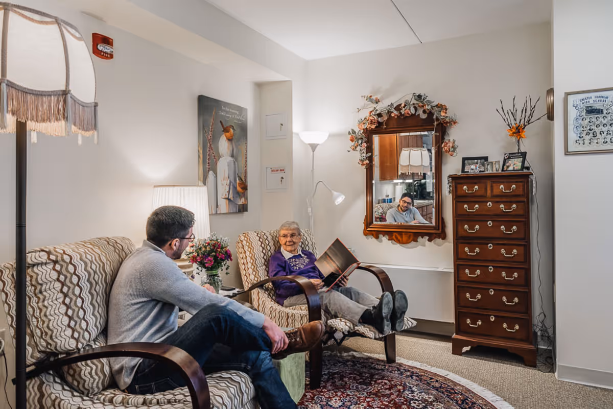 An elderly woman and a man sit and chat in a cozy living room area with patterned armchairs, lamps, a mirror and a wooden dresser.