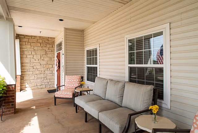 Covered outdoor porch area with a cushioned sofa, two cushioned chairs, small tables with yellow flowers in vases, beige siding walls, two windows with white frames, and a stone wall near the entrance door.