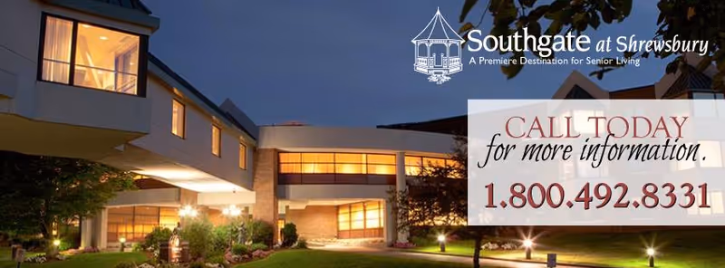 Exterior view of Southgate at Shrewsbury senior living facility at dusk, showing a modern building with large windows illuminated from inside, surrounded by landscaped greenery and outdoor lighting.