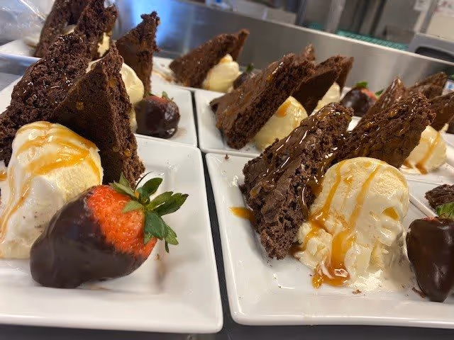 Close-up view of dessert plates featuring scoops of vanilla ice cream drizzled with caramel sauce, triangular pieces of chocolate brownie, and chocolate-covered strawberries arranged neatly on white square plates.
