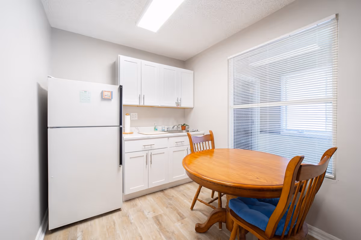 A small kitchen area with white cabinets, a white refrigerator, a countertop with a sink, and a wooden dining table with two wooden chairs that have blue cushions. There is a large window with blinds on the right side, and the floor has light-colored wood flooring.