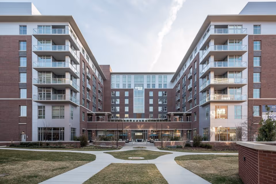 Exterior view of a modern multi-story senior living facility with brick and glass facade, balconies, and a landscaped courtyard with paved walkways leading to the entrance.