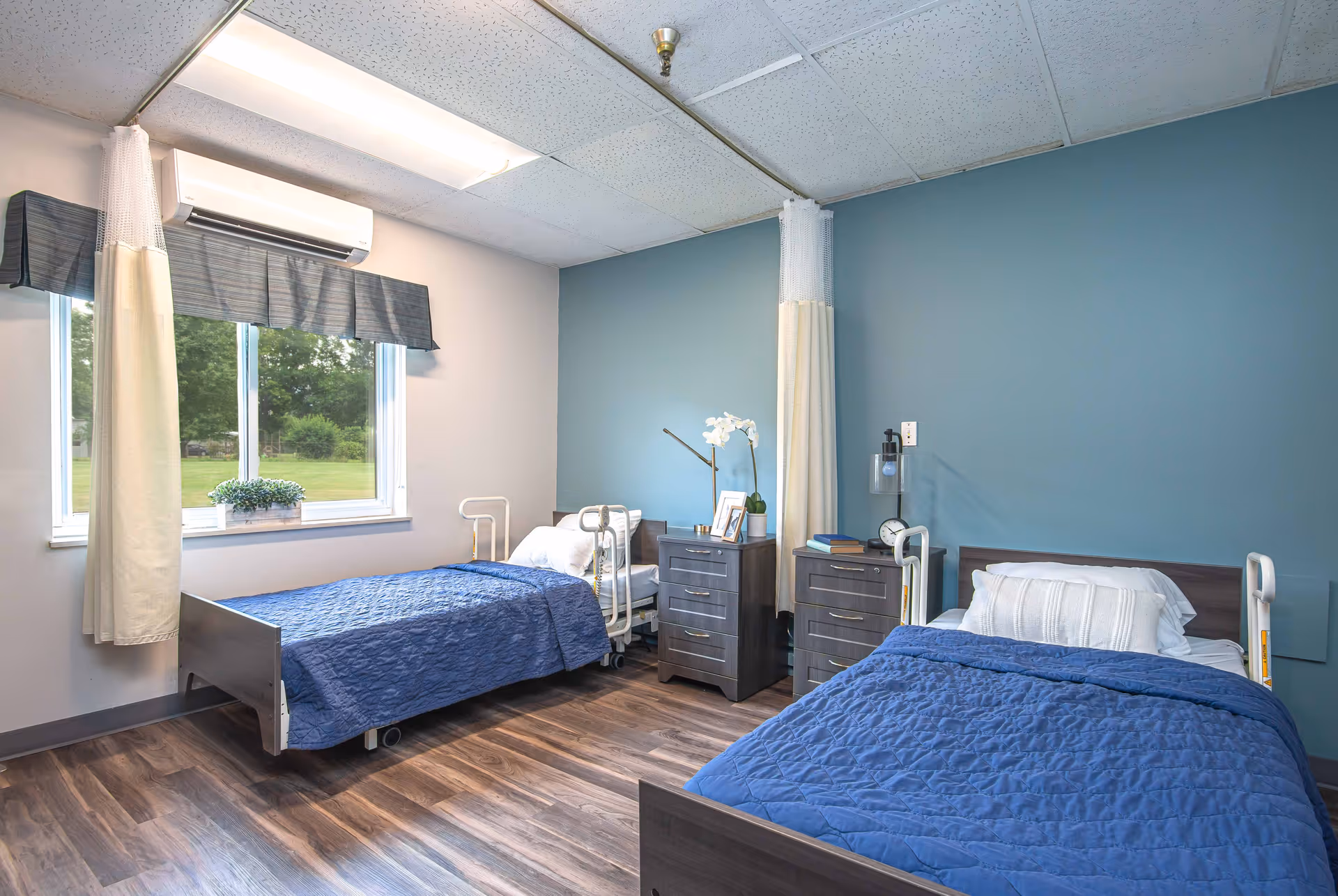 A bright and clean bedroom in a senior living facility with two single beds, each with blue quilts and white pillows. Between the beds are two dark wooden nightstands with a lamp, clock, books, and a small plant. A large window with curtains lets in natural light, and the walls are painted light blue and white. The floor is wood laminate.