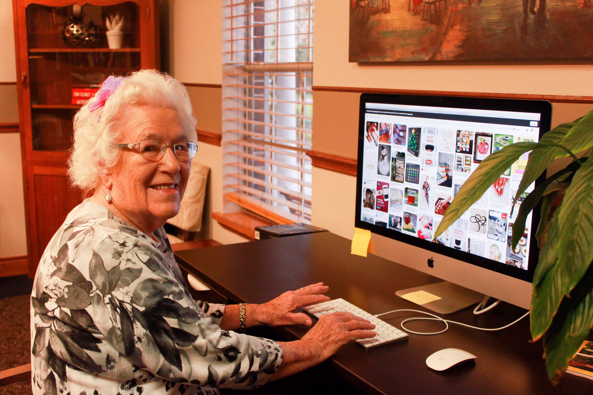 An elderly woman with white hair and glasses is sitting at a desk using a desktop computer. She is smiling and looking towards the camera. The room has wooden furniture, a window with blinds, a plant on the desk, and a painting on the wall.