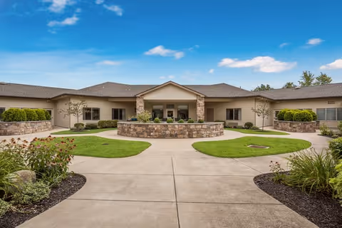 Exterior view of a single-story senior living facility building with a central entrance, stone accents, and landscaped garden beds with green shrubs and flowers under a blue sky with scattered clouds.