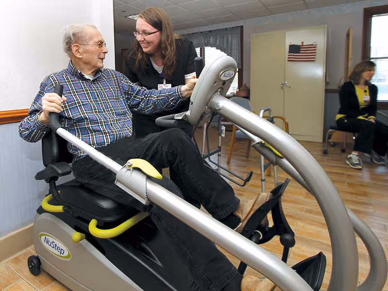 An elderly man using a NuStep exercise machine in a healthcare facility while a female staff member smiles and interacts with him. Another woman is seated in the background near a cabinet with an American flag on it. The room has wooden flooring and light-colored walls.