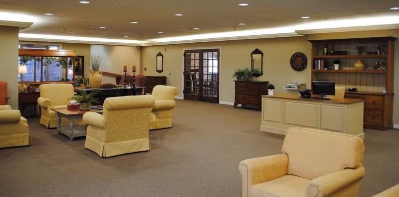 A spacious and well-lit common area in a senior living facility with beige carpet and walls. The room features several upholstered armchairs arranged around a coffee table with a plant centerpiece. In the background, there is a wooden reception desk with a computer and printer, a wooden cabinet with shelves holding decorative items, and a set of double glass doors. Additional furniture includes a chest of drawers with a plant and a wall clock above it, as well as mirrors and candle holders on the walls.