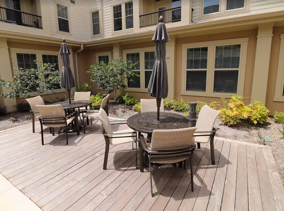 Outdoor patio area with two round metal tables, each surrounded by four beige cushioned chairs. Each table has a closed dark umbrella in the center. The patio is made of wooden planks and is adjacent to a beige building with multiple windows and some greenery along the building's edge.