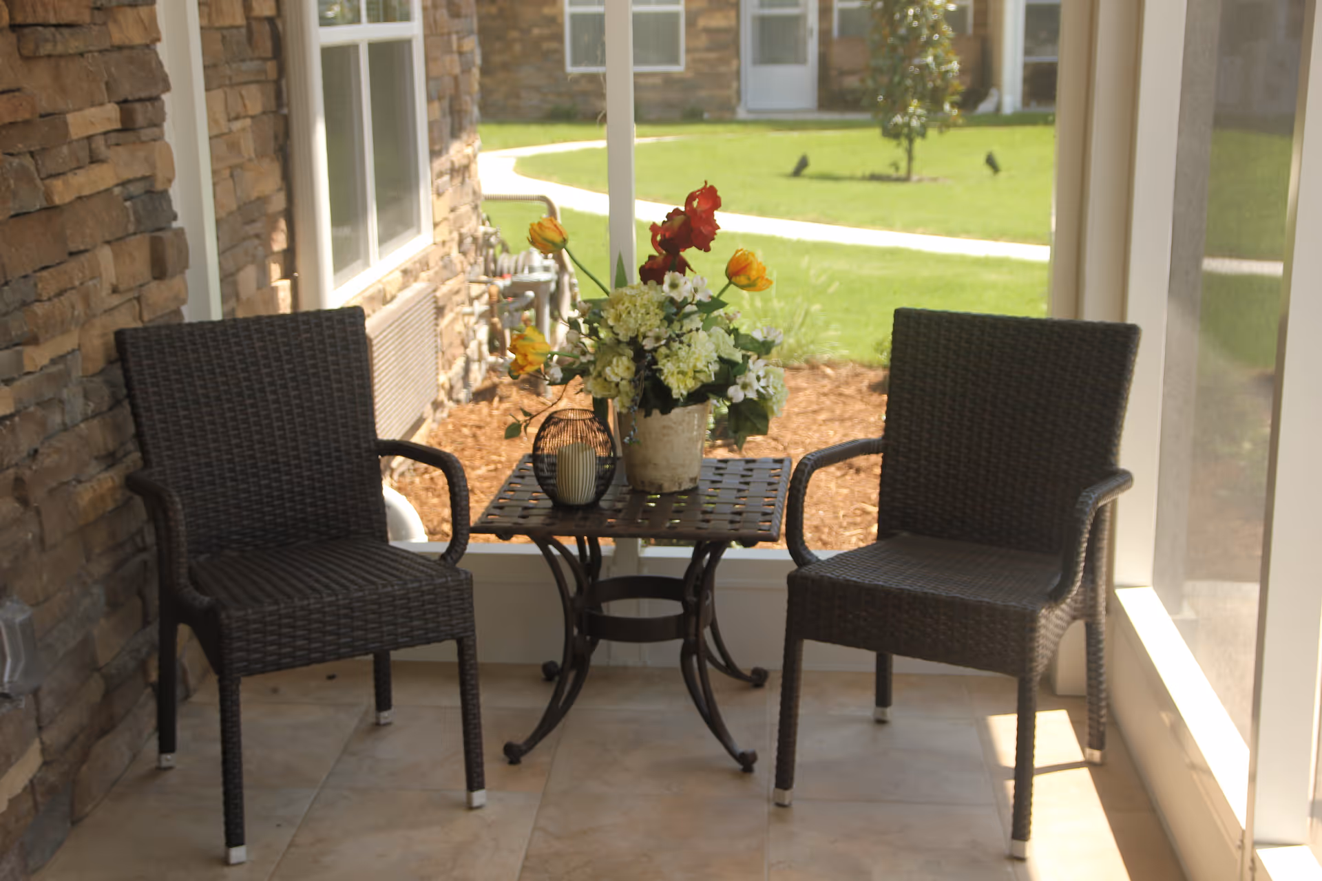Two dark wicker chairs with armrests are placed on either side of a small square metal table with a lattice design. On the table, there is a flower arrangement with white, yellow, and red flowers, along with a decorative candle holder. The seating area is on a tiled floor next to a stone wall and large windows, overlooking a green lawn with a curved pathway and some small trees.