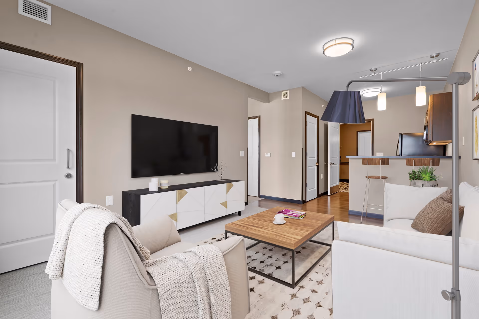 A modern living room with a beige armchair draped with a knitted blanket, a white sofa with a brown cushion, a wooden coffee table with a cup and a magazine, a large flat-screen TV mounted on the wall above a white and black TV stand, and a kitchen area with bar stools and pendant lights in the background.