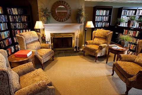 A cozy living room with four upholstered armchairs arranged around a fireplace. The fireplace has a white mantel with a round mirror above it and two plants on either side. There are bookshelves filled with books along the walls and two table lamps providing warm lighting.