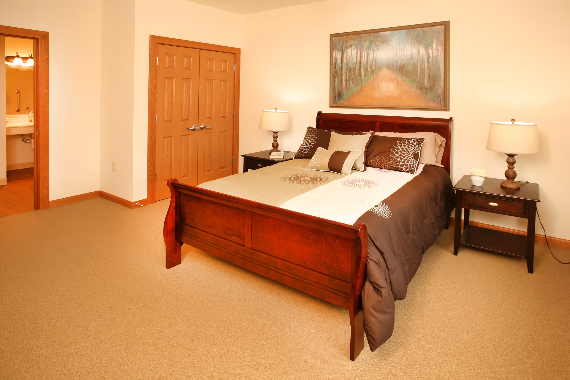 A bedroom with a wooden bed frame and a neatly made bed with brown and beige bedding. There are two matching nightstands on either side of the bed, each with a lamp. A painting of a forest path hangs on the wall above the bed. A double-door closet and an open door leading to a bathroom are visible in the background.
