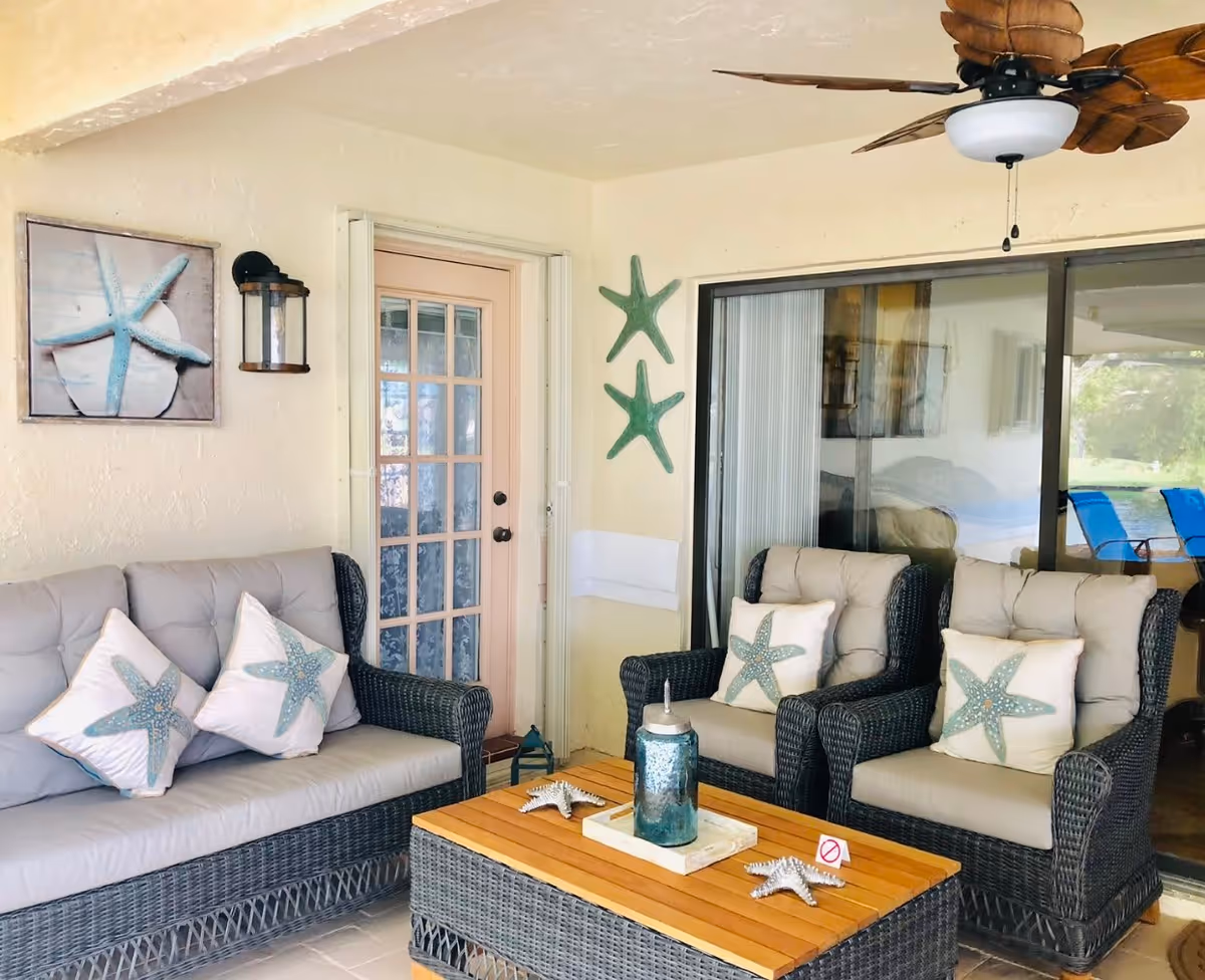 Covered patio seating area with wicker sofa and armchairs, starfish-themed pillows, a wooden coffee table and a ceiling fan.
