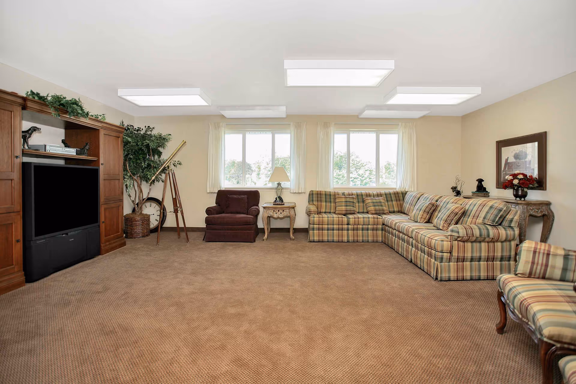 Spacious communal living room with a plaid sectional sofa, armchairs, an entertainment center and windows.