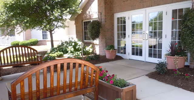 Outdoor seating area with wooden benches and flower planters in front of a brick building with white double glass doors. There is a tree and various plants around the walkway leading to the entrance.
