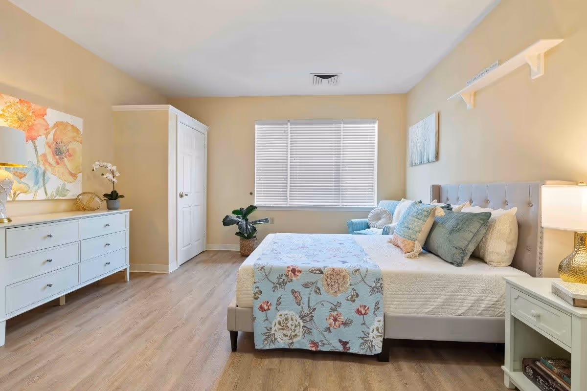 A bright and cozy bedroom with a large bed adorned with multiple pillows and a floral bed runner. The room features light wood flooring, a white dresser with a lamp and decorative items, a potted plant near a window with blinds, a blue armchair, and soft beige walls with floral artwork.