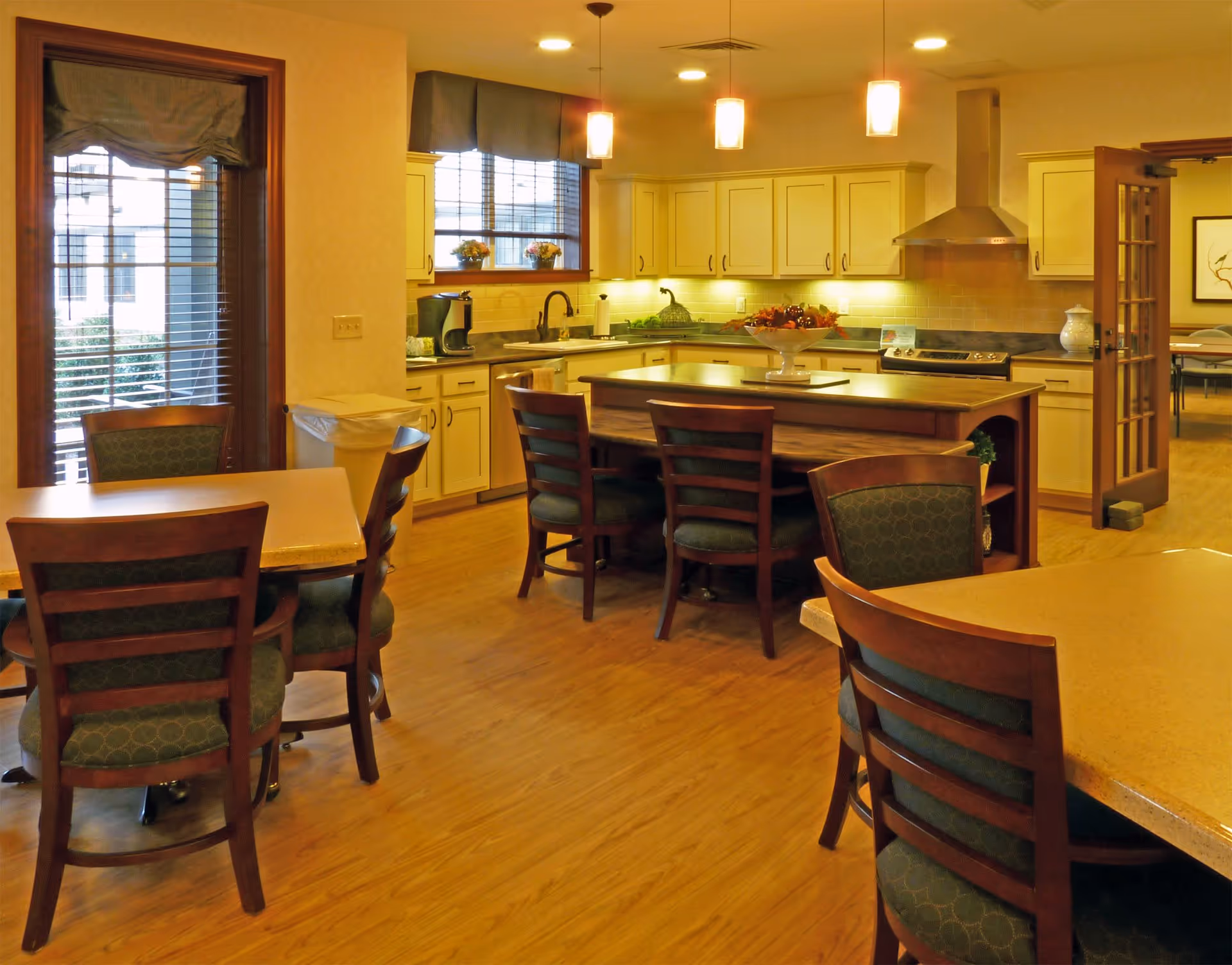 A warm, well-lit kitchen and dining area with wooden floors, cream-colored cabinets, a kitchen island with chairs, pendant lights, and tables with chairs. There is a window with blinds and a door with glass panels leading to another room.