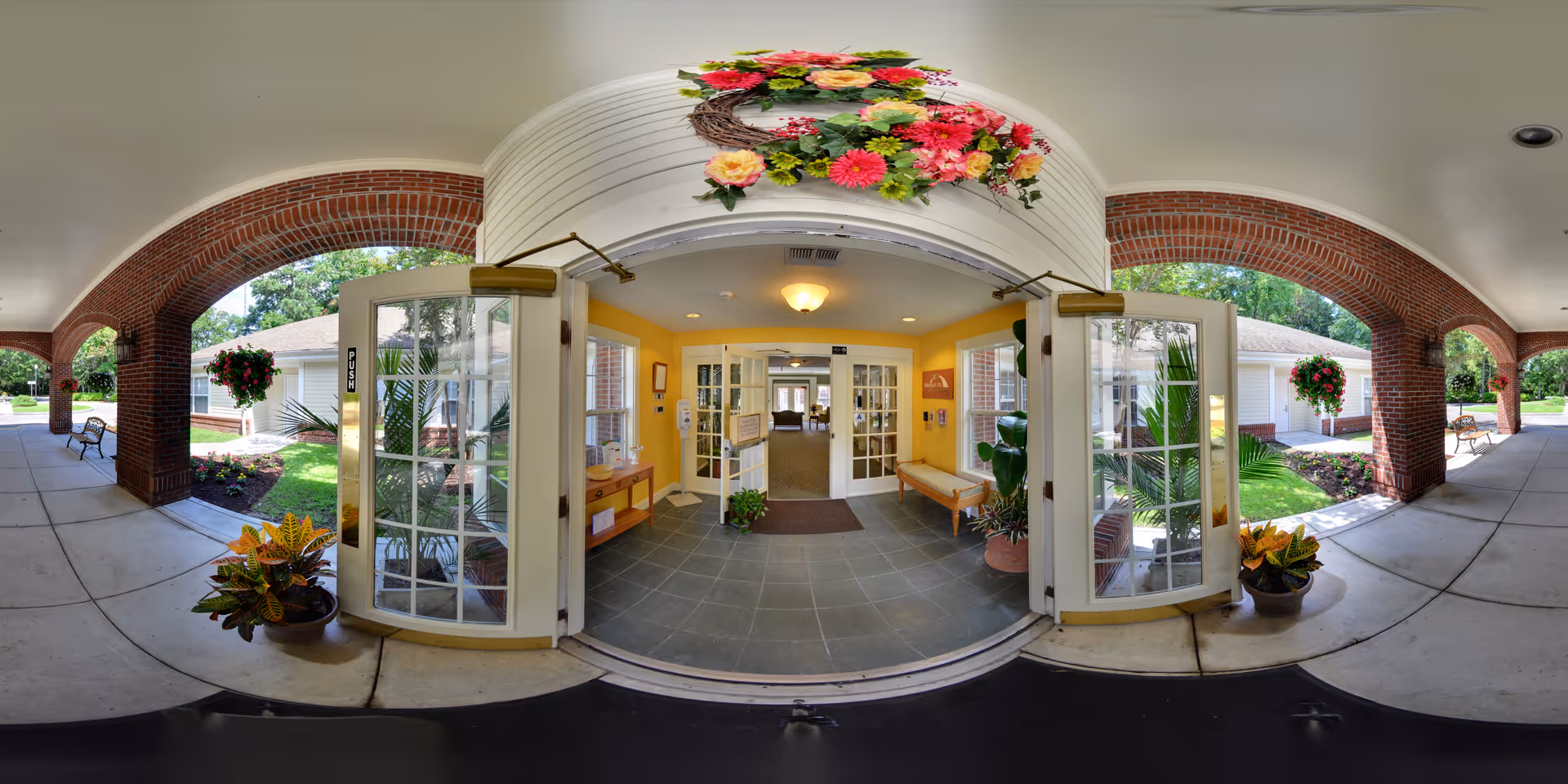 Entrance to a senior living facility with open double glass doors leading into a lobby area. The exterior features brick archways and a covered walkway with benches and potted plants. Above the entrance is a decorative floral wreath with colorful flowers. The interior lobby has yellow walls, a bench, plants, and a tiled floor.