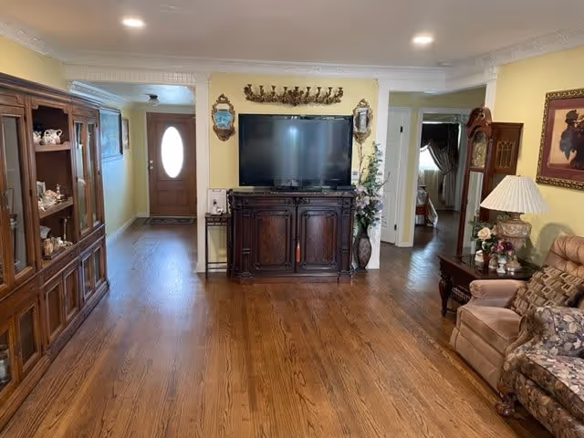 Bright living room with hardwood floors, a large TV on a wooden cabinet centered on a yellow wall, display cabinets and upholstered seating.