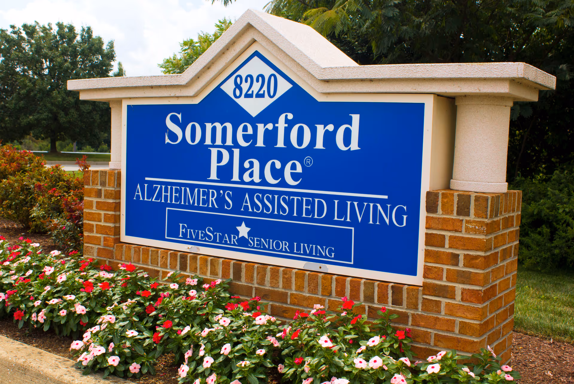 Outdoor blue sign for Somerford Place Alzheimer's Assisted Living (8220) mounted on a brick base with flowers planted in front.