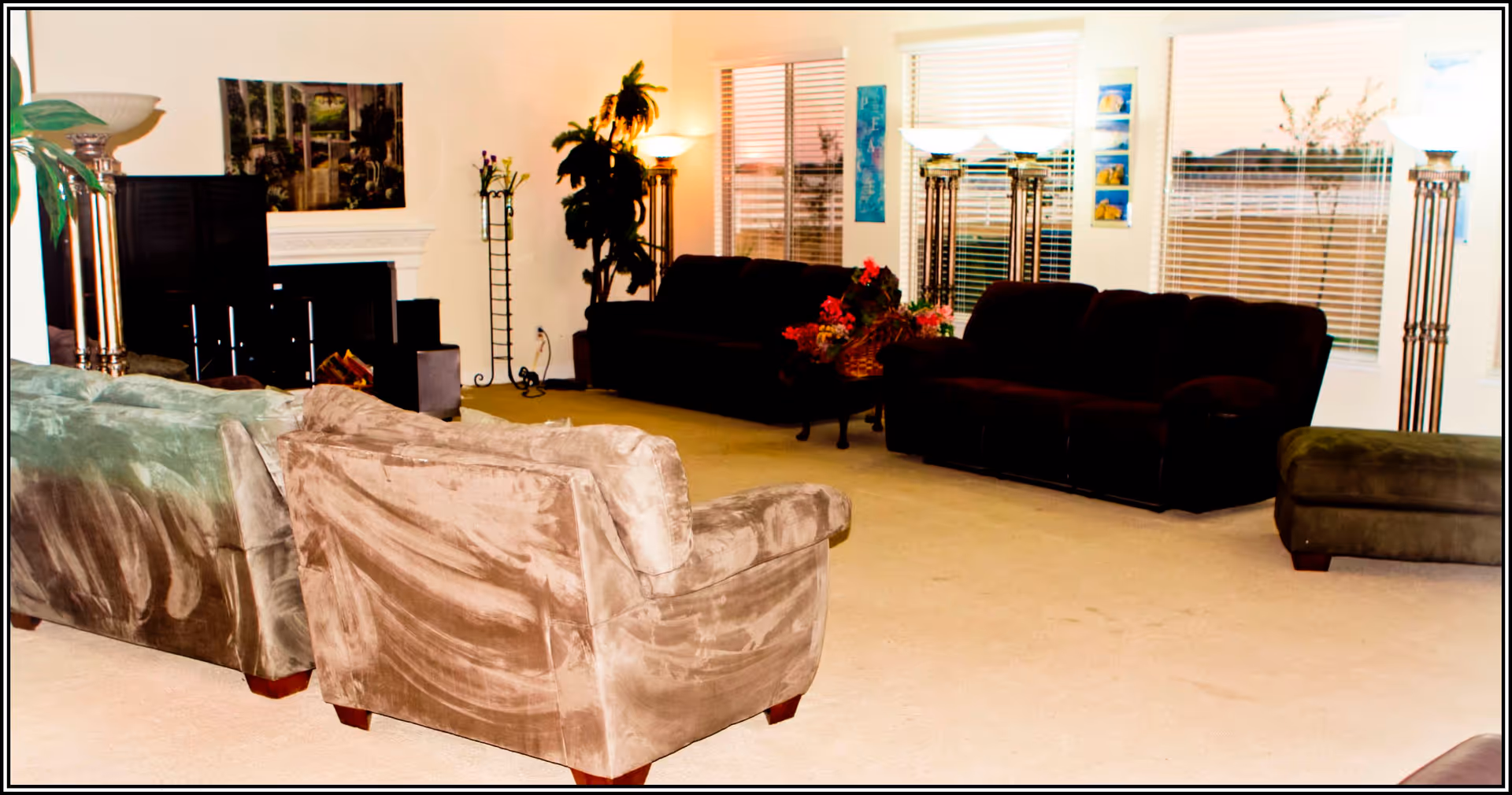 A spacious living room with multiple sofas and armchairs arranged around a central area. There are large windows with blinds letting in natural light, a fireplace with a painting above it, tall floor lamps, and some indoor plants and floral decorations.