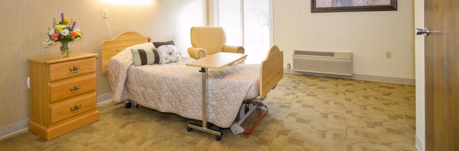 A furnished single-occupancy bedroom with an adjustable bed, wooden nightstand with flowers, armchair, and overbed table.
